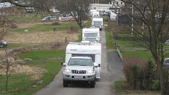 En bil trækker en campingvogn på en grusvej, omgivet af flere campingvogne og et grønt landskab.