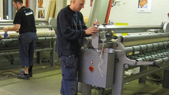 En mand arbejder ved en maskine i en fabrik, mens en anden mand står i baggrunden.
