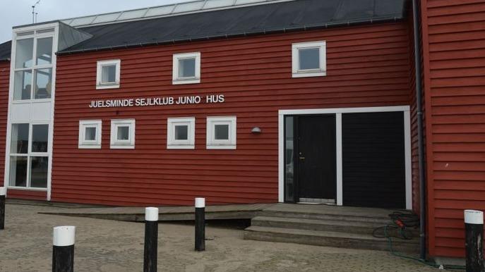 Rød bygning med tekst "Jelsminde Sejlklub Juno" og sorte døre, beliggende ved en parkeringsplads.