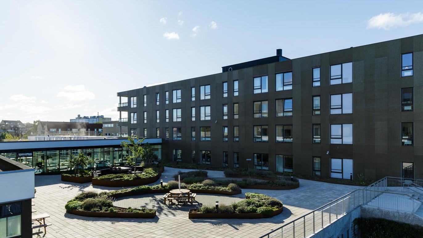 Moderne bygning med flere etager og en indbydende gårdhave med grønne planter og siddepladser.