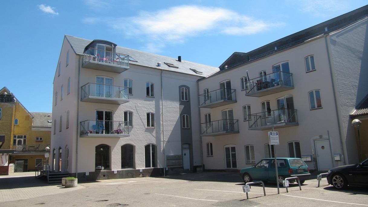 Moderne lejlighedsbygning med altaner og blå himmel i baggrunden. Parkeringsplads foran bygningen.