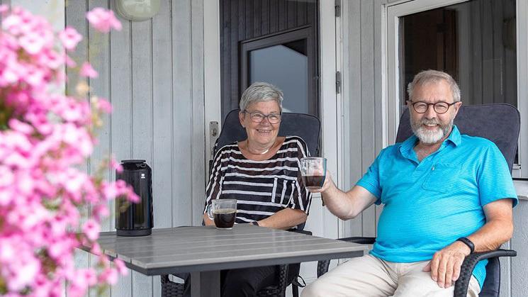 To personer sidder på en terrasse med drinks, omgivet af blomster i en hyggelig atmosfære.