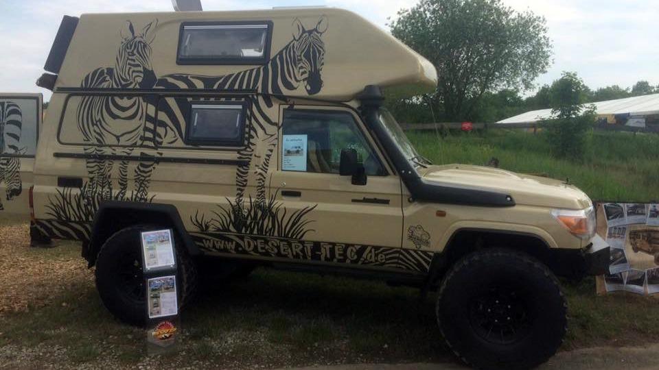 En camper med zebra-motiv står parkeret på en grusvej, omgivet af grønne områder og udstillingsmateriale.