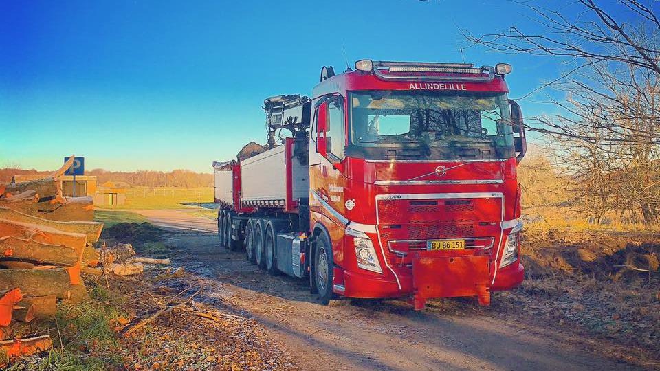 En rød lastbil parkeret på en grusvej med træstammer i baggrunden og en klar blå himmel.