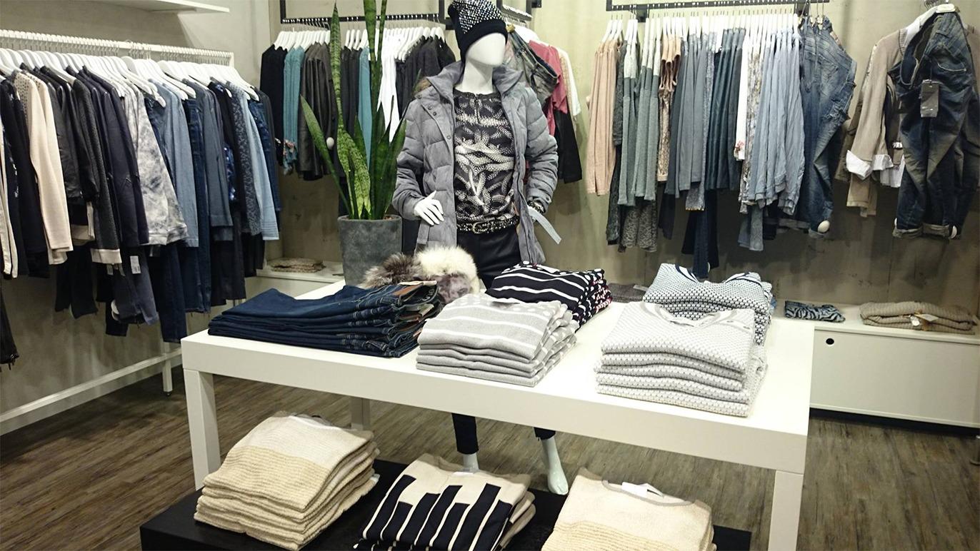 Butik med tøj, herunder en mannequin iført en jakke, og stablede trøjer og bukser på et bord.