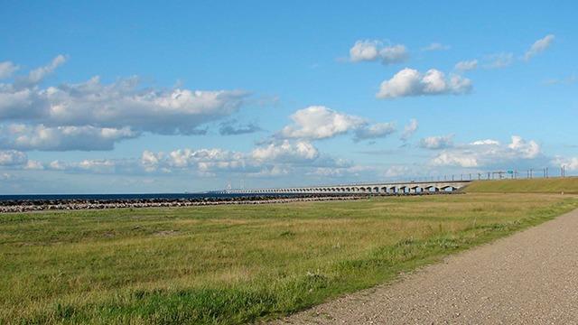 En græsmark med en grusvej, der fører til en dæmning under en blå himmel med skyer.