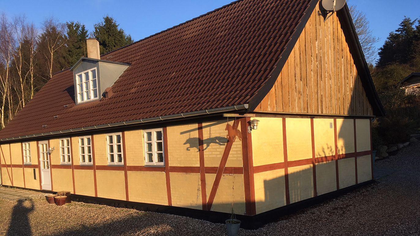 Et stort hus med træbeklædning og rødt tag, beliggende i et naturskønt område med sollys.