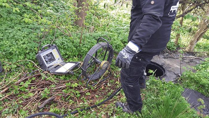 En person i arbejdstøj står ved en computer og en spole i en grøn have med buske og græs.