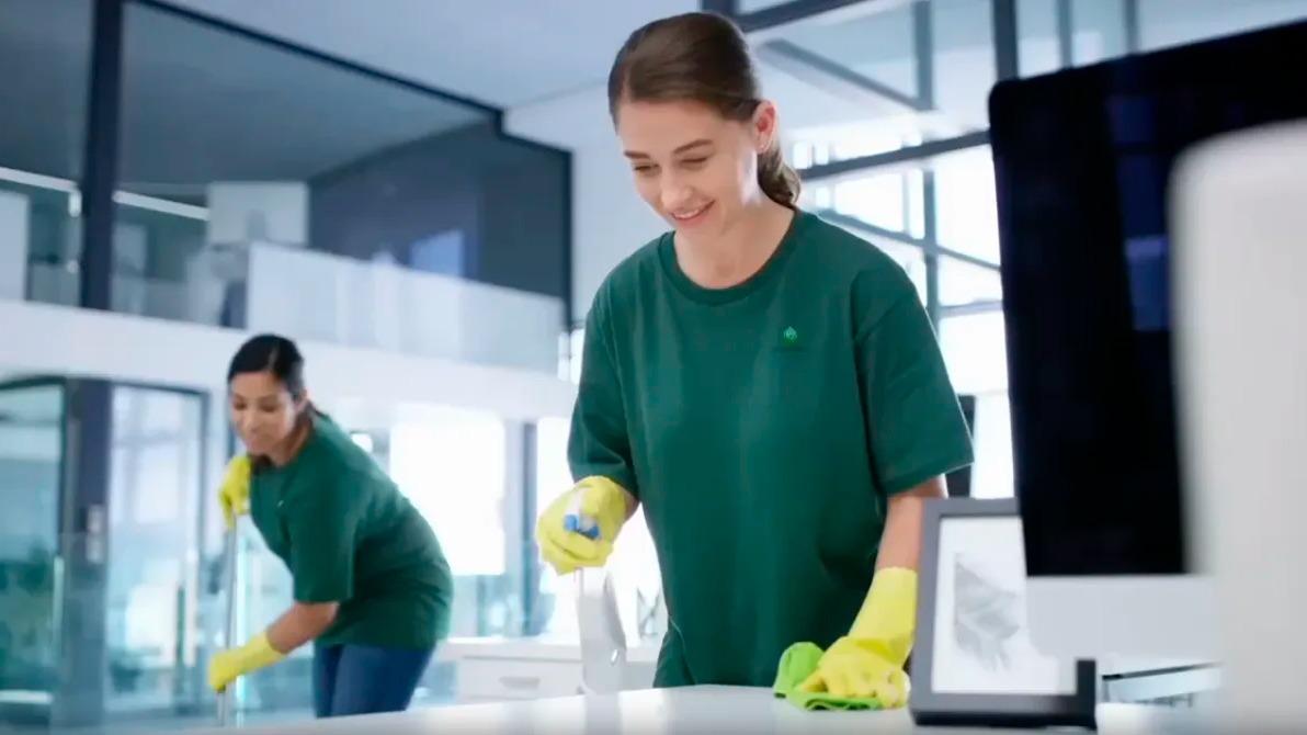 To kvinder, der gør rent i et moderne kontor, iført grønne t-shirts og gule handsker, mens de rengør borde.