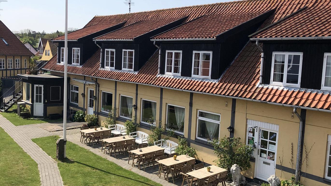 En bygning med flere etager og rødt tag, omgivet af borde og stole på en udendørs terrasse.