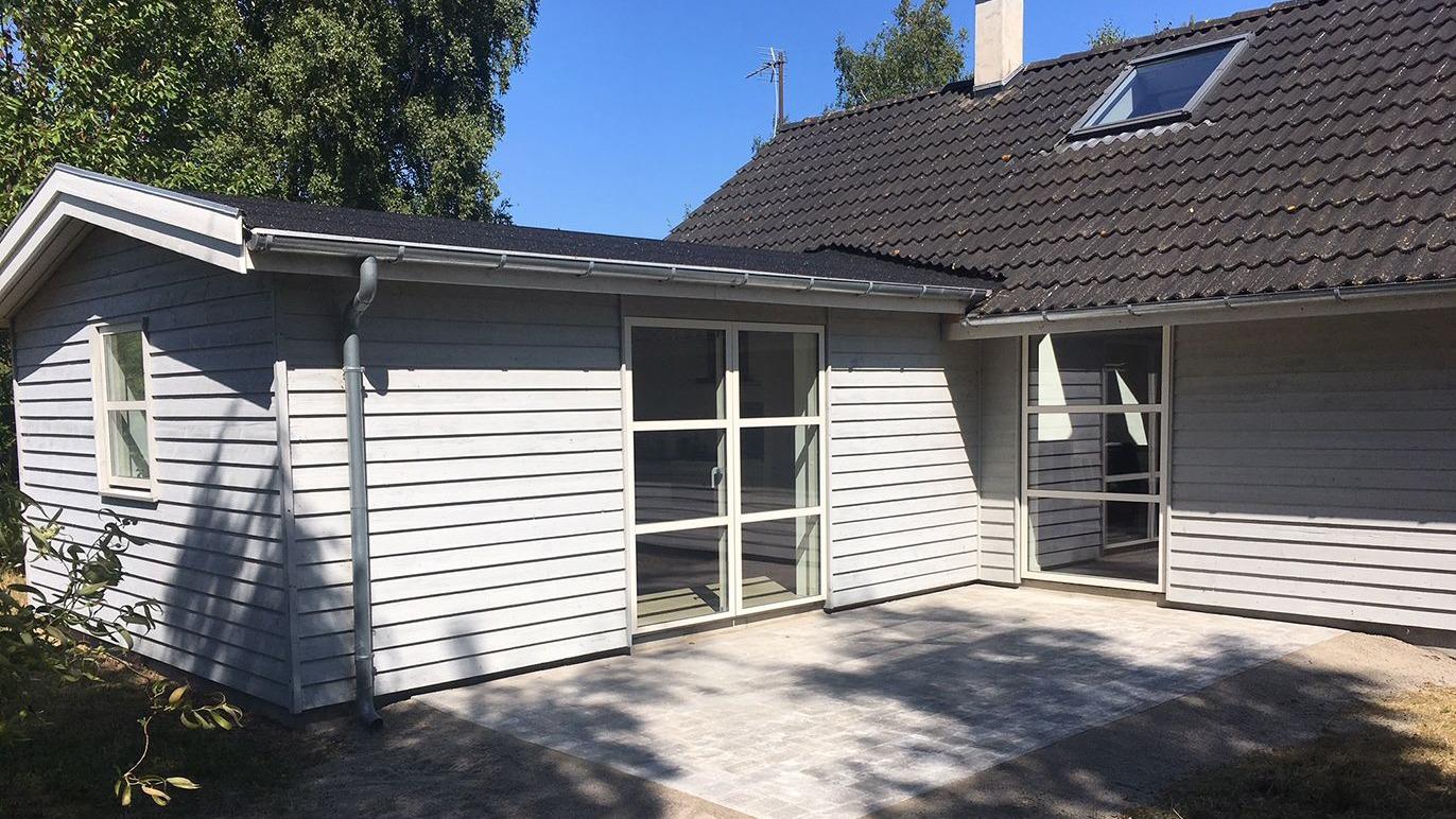 Moderne grå træhus med fladt tag og store vinduer, beliggende i en solrig have.