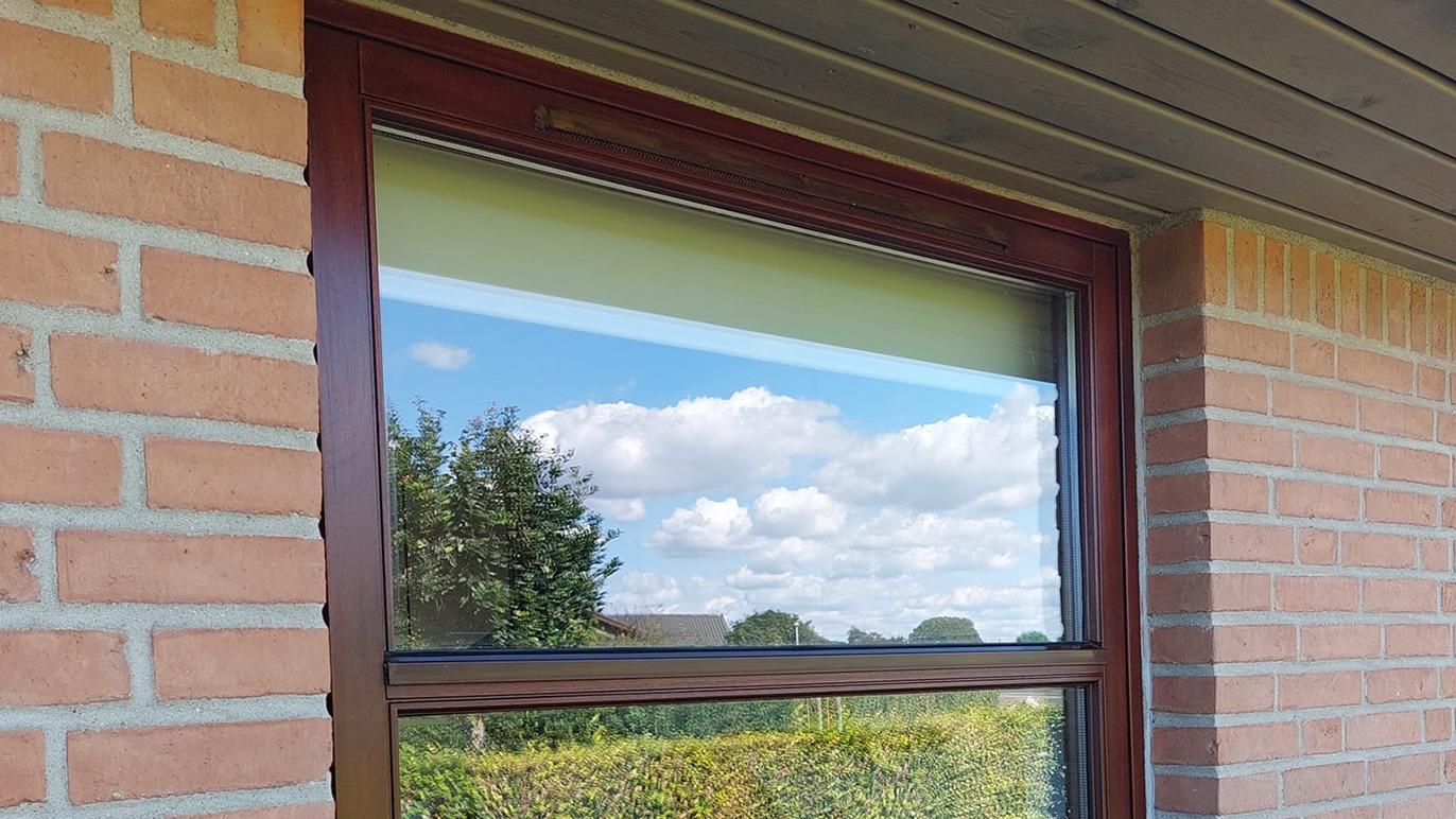 Vindue med brune rammer, der reflekterer en blå himmel og skyer, set fra en murstensvæg.