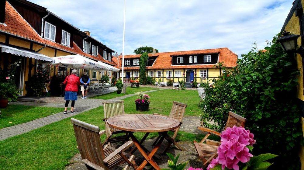 En hyggelig gårdhave med træmøbler, blomster og bygninger med røde tage under en skyet himmel.