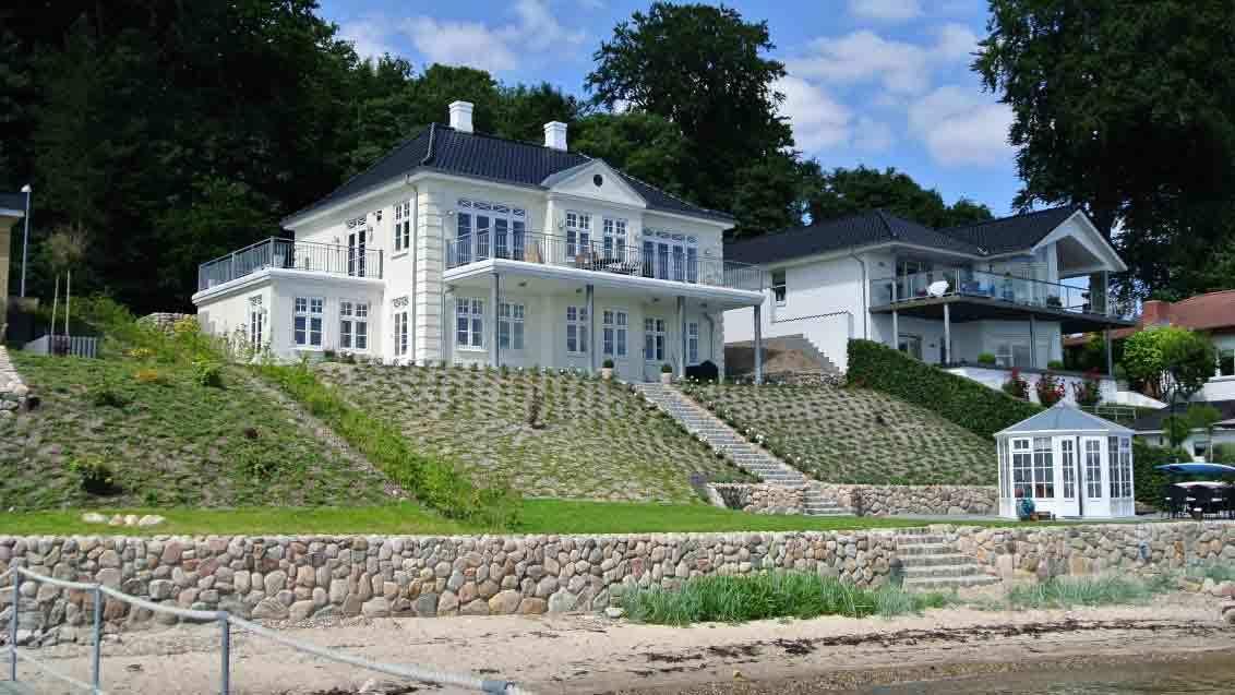 Et smukt hvidt hus ved vandet med en stor terrasse og grønne skråninger omkring.