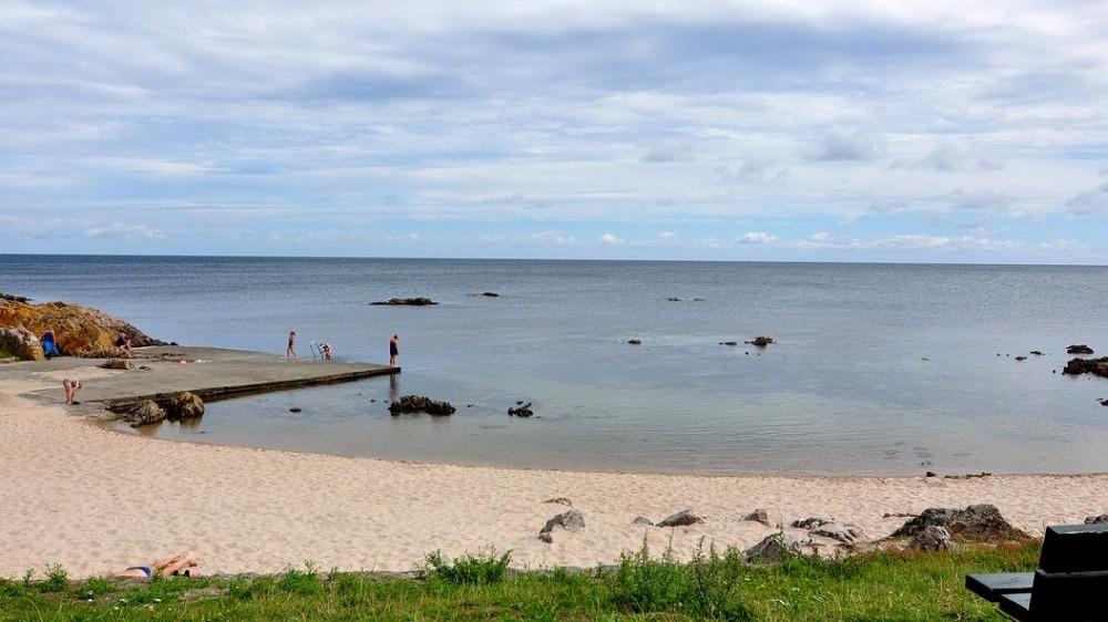 En strand med sand, en mole og mennesker, der står ved vandkanten under en skyet himmel.