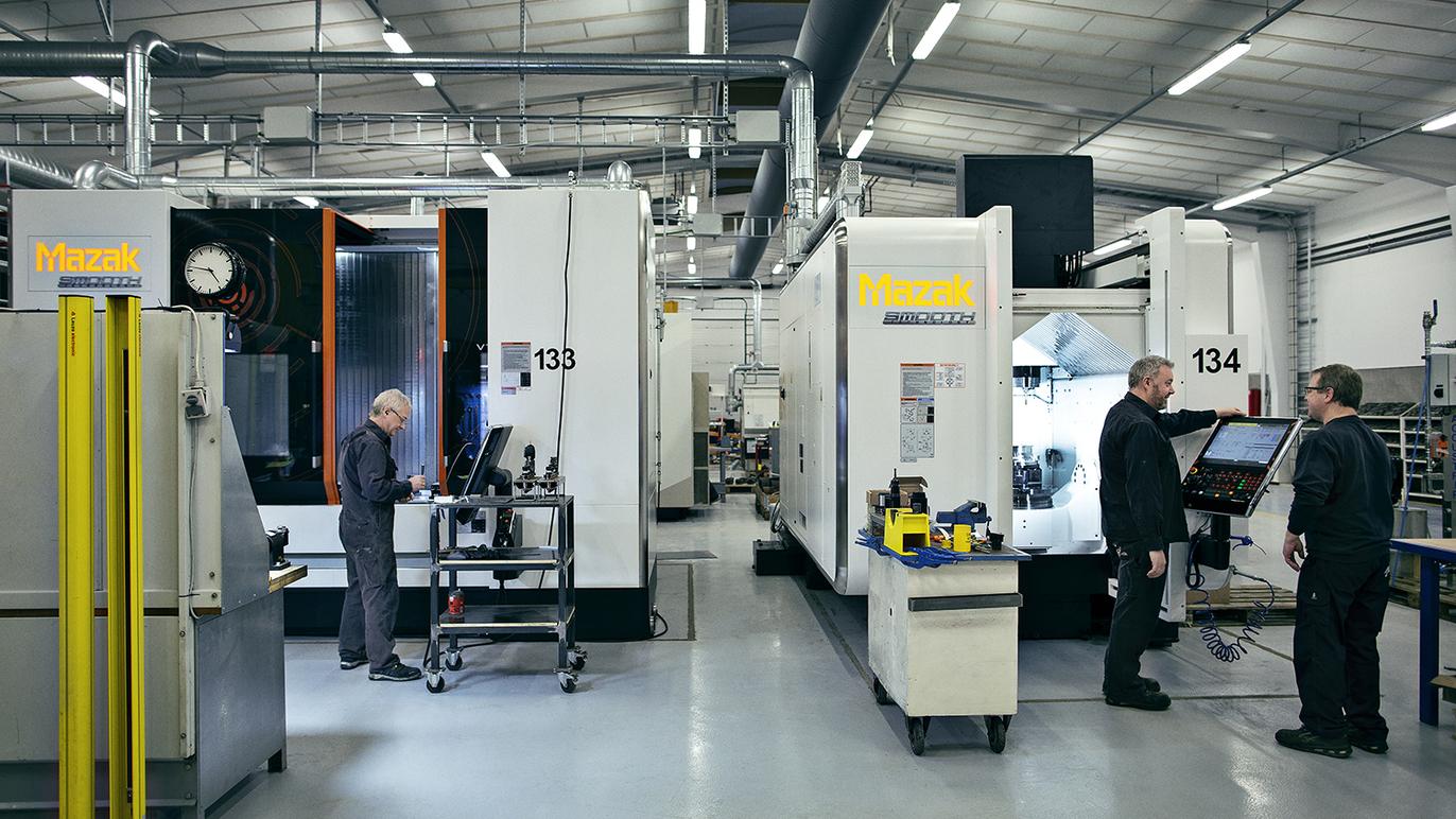 Moderne produktionshal med maskiner og teknikere, der arbejder ved CNC-maskiner og overvåger processer.