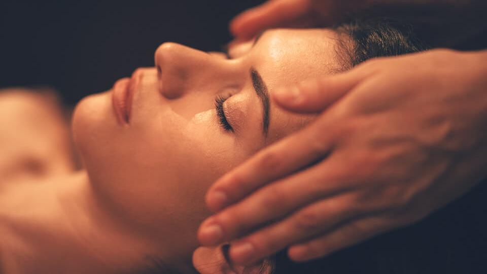 Kvinde får ansigtsmassage med hænderne, afslappet ansigtsudtryk og lukkede øjne i et roligt miljø.