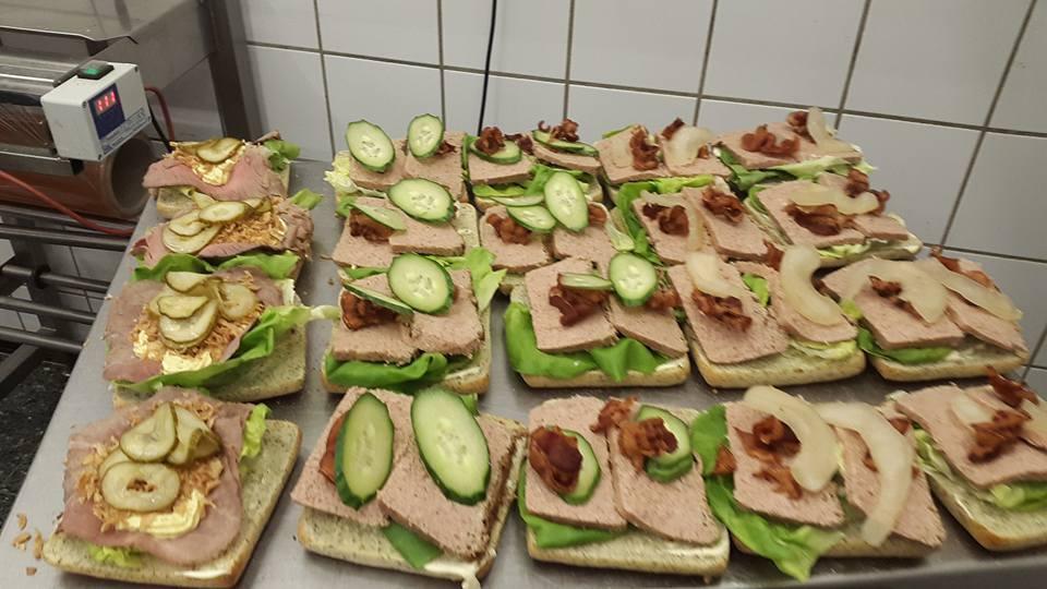 Flere smørrebrød med leverpostej, agurk, bacon og salat på en metalplade i et køkken.