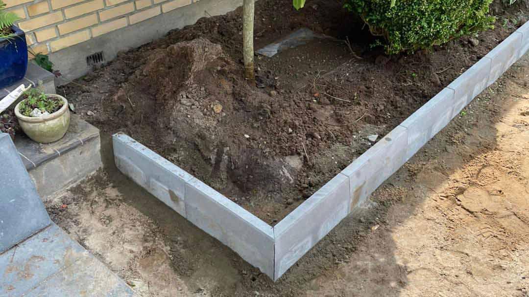 Et nyanlagt bed med en betonkant, en lille busk og en potteplante ved siden af. Jorden er løs og uformet.
