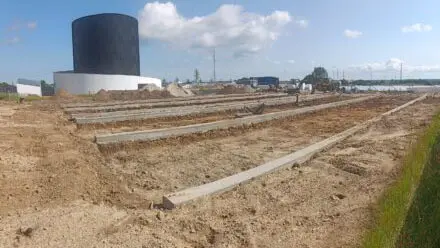 Byggeplads med udgravede områder og betonfundamenter, en stor sort tank i baggrunden under en blå himmel.