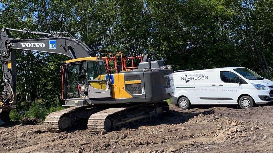 En stor Volvo gravemaskine står på en byggeplads ved siden af en hvid varevogn.