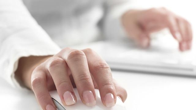 Hånd der bruger en computer mus, med en anden hånd på et tastatur, i et lyst kontormiljø.