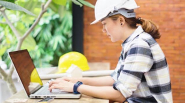 En kvinde i hjelm arbejder på en bærbar computer i et byggeområde med gule hjelme i baggrunden.