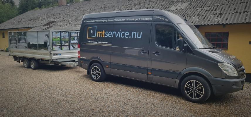 En grå varevogn med en trailer parkeret foran en bygning med tag af tagpap.