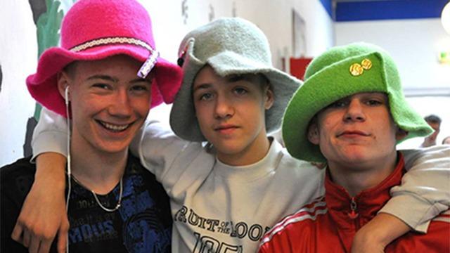 Tre unge mænd står sammen og smiler, iført farverige hatte i pink, grøn og grå.
