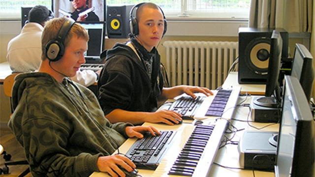 To unge mænd sidder ved computere med hovedtelefoner og arbejder med musikproduktion i et klasseværelse.