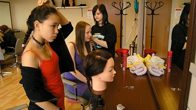 To unge piger i en frisørsalon, der arbejder med en hovedskulptur og ser sig selv i spejlet.
