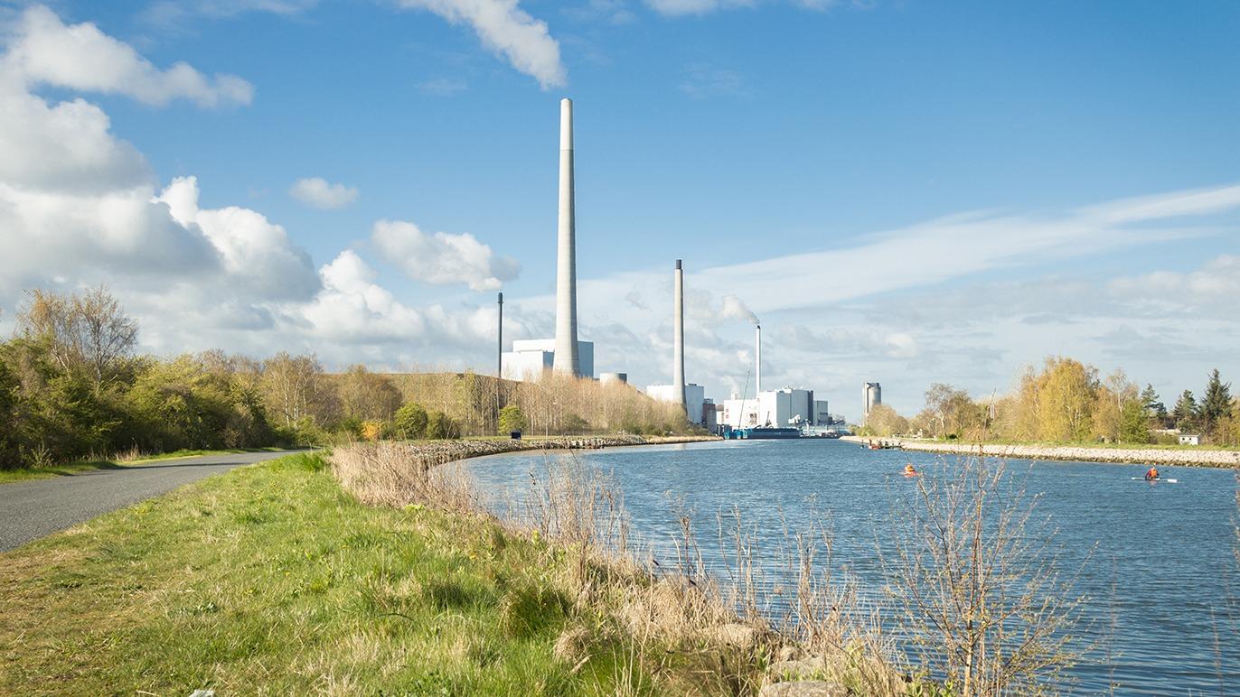 En industriel fabrik ved en sø med grønne områder og en blå himmel med skyer.