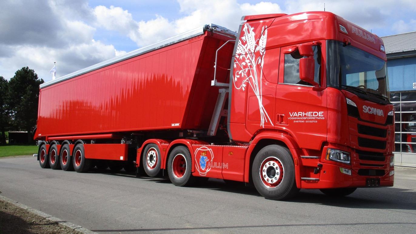 En rød lastbil med en stor trailer parkeret på en grusvej ved en bygning.
