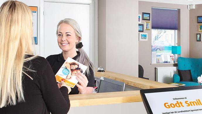En smilende receptionist giver en kunde en brochure ved skranken i en moderne reception.