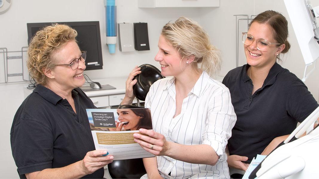 Tre kvinder i en tandklinik, hvor de ser på en brochure og smiler, mens de diskuterer tandpleje.
