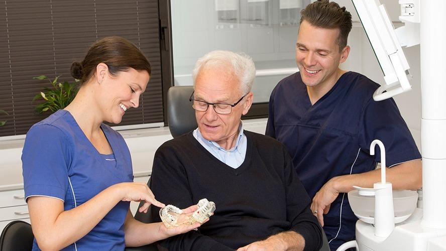 En tandlægeassistent viser en ældre mand en tandmodel, mens en tandlæge smiler i baggrunden.