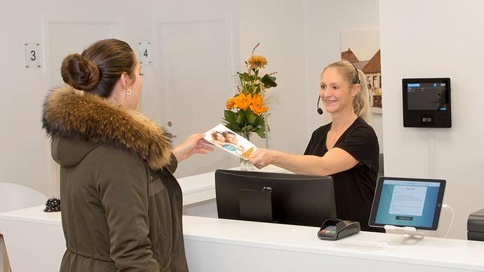 En kvinde i en jakke modtager en brochure fra en smilende receptionist ved en skranke.