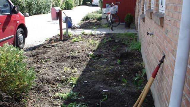 En nyanlagt have med jord, en skovl og en rød bil parkeret ved siden af. En cykel står også i baggrunden.