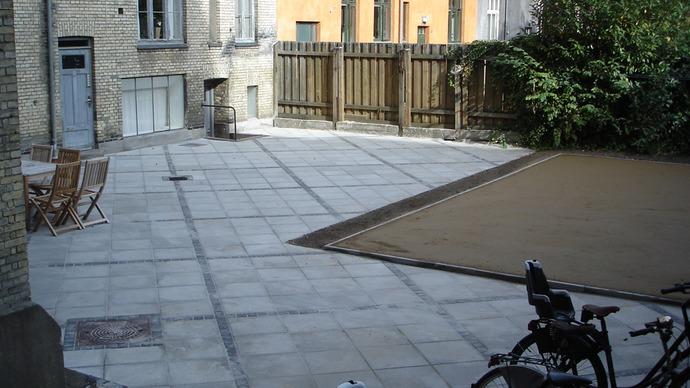 En gård med fliser, en cykel og et træhegn i baggrunden. Der er også en lille terrasse med havemøbler.