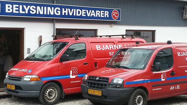 To biler fra Bjarne Lund med logo og skilt for Belysning Hvidevarer i baggrunden.