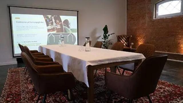 Mødelokale med et langt bord, stole, projektor og præsentation på væggen. Indretningen er moderne og indbydende.