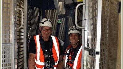 To mænd iført sikkerhedshjelme og refleksveste står i en elevator, klar til arbejde i en industrielt miljø.