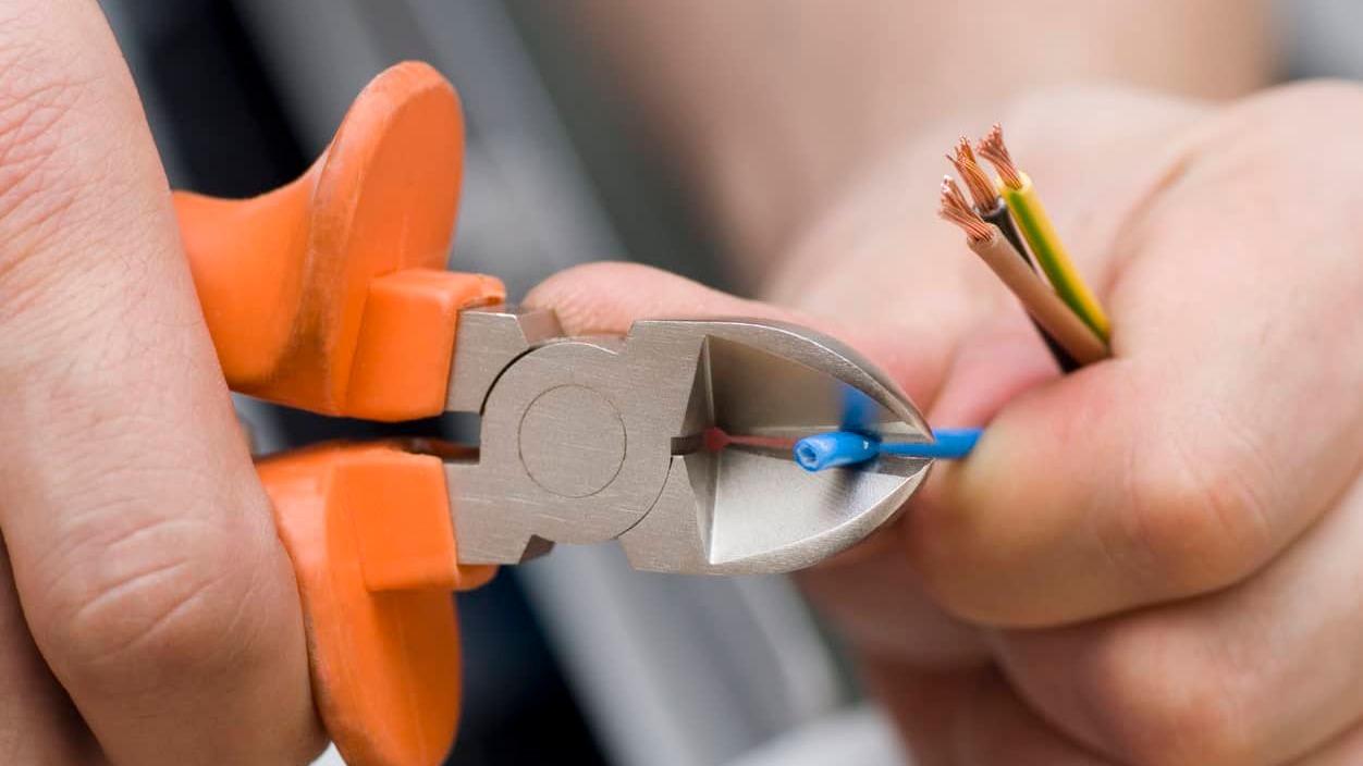 Hånd, der holder en afisoleringstang, mens den klipper elektriske ledninger i forskellige farver.