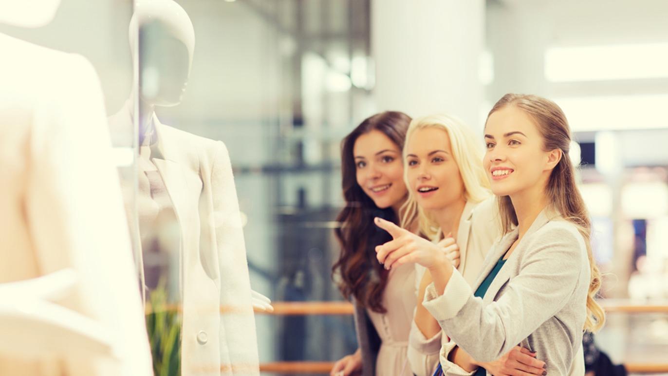 Tre kvinder kigger på et vindue med en mannequin iført en hvid dragt i et moderne shoppingcenter.