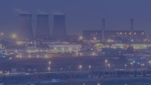 Aftenscene med industrielle bygninger og køletårne oplyst af byens lys.