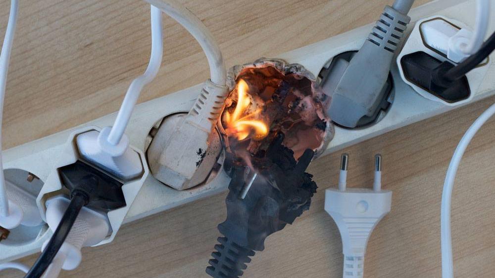 Brændende stik i en stikkontakt med røg og flammer, omgivet af flere ledninger og stik.