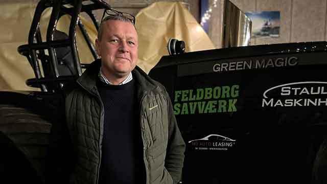 Mand står foran en stor traktor med teksten "FELDBORG SAVVÆRK" og "GREEN MAGIC" i en hal.