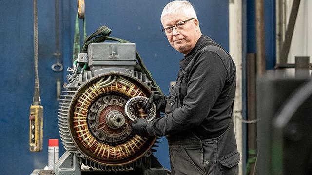 En mand i arbejdstøj står ved en stor motor og holder en del af motoren i hånden.