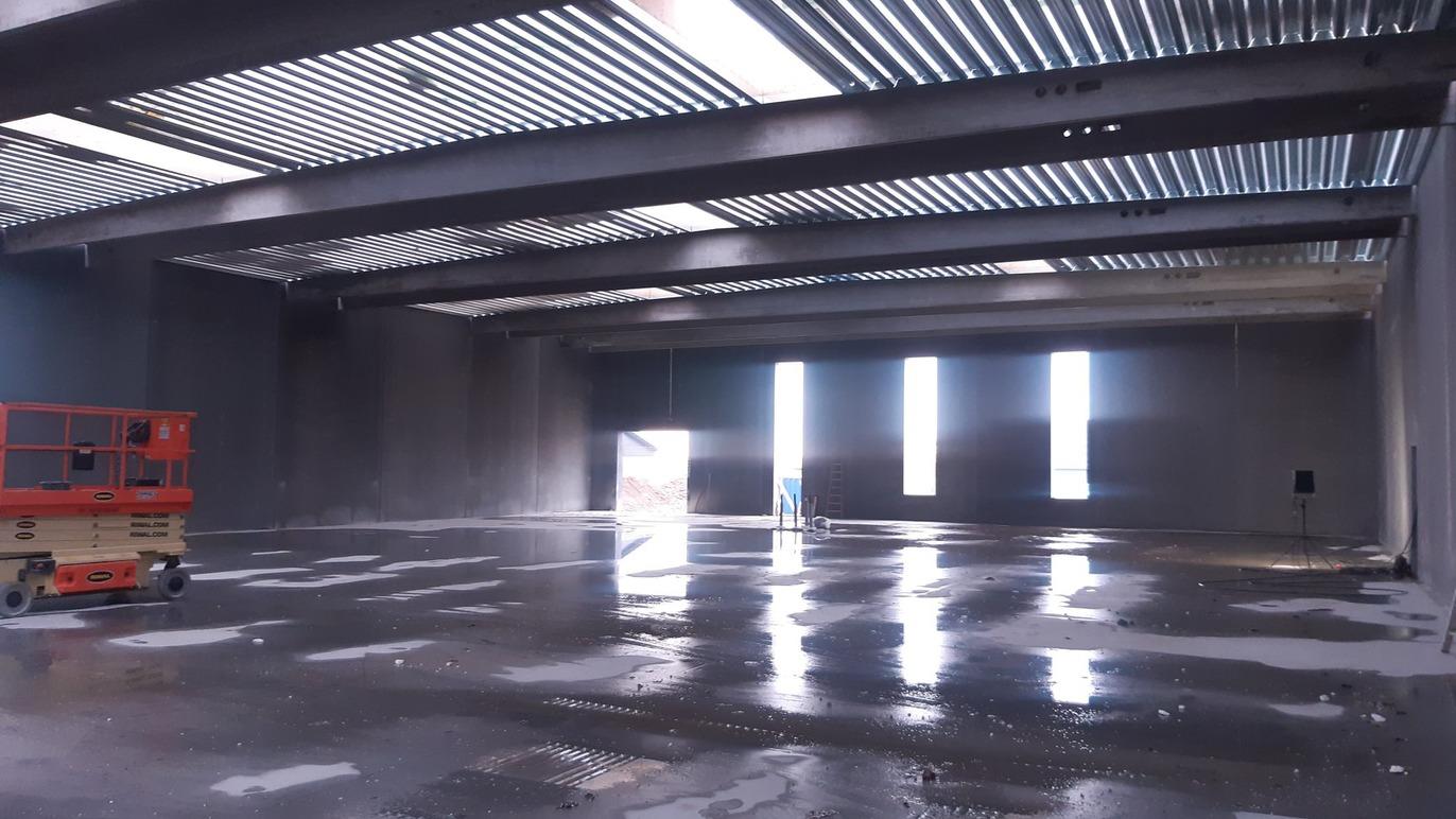 Tom lagerhal med metalbjælker, store vinduer og vådt gulv, der reflekterer lyset.