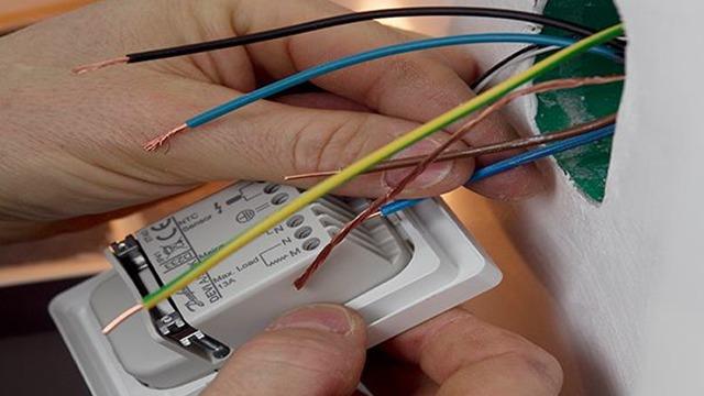 Hånd, der tilslutter ledninger til en elektrisk kontakt i en væg, med synlige farvede ledninger.